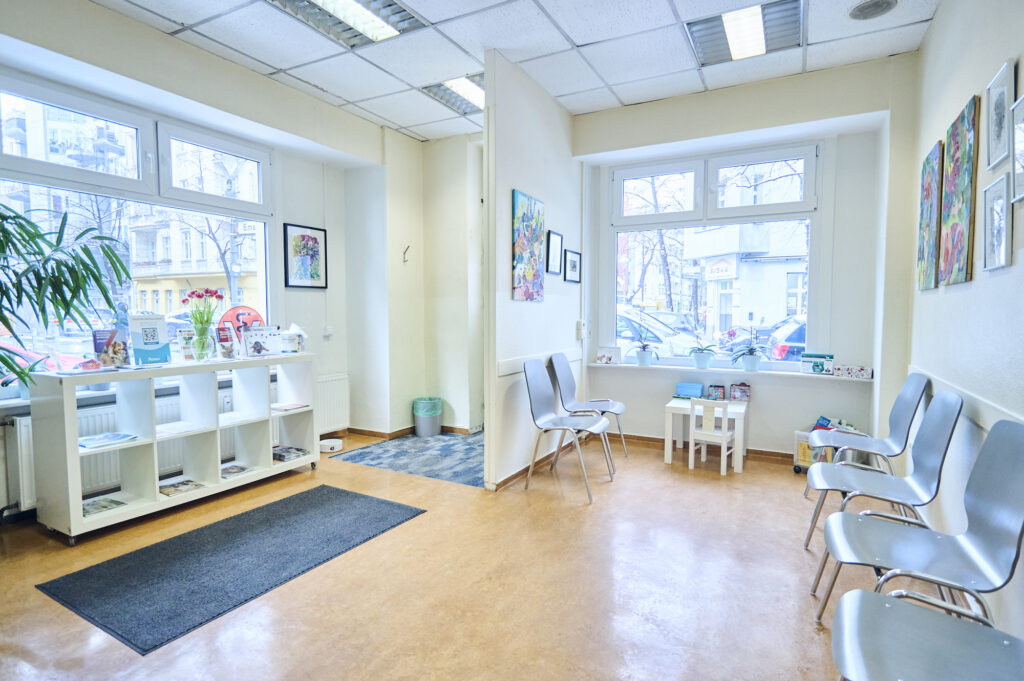 Helles Wartezimmer einer Tierarztpraxis mit Stühlen, kleiner Spielecke, Regal, großen Fenstern und dekorativen Bildern an den Wänden.