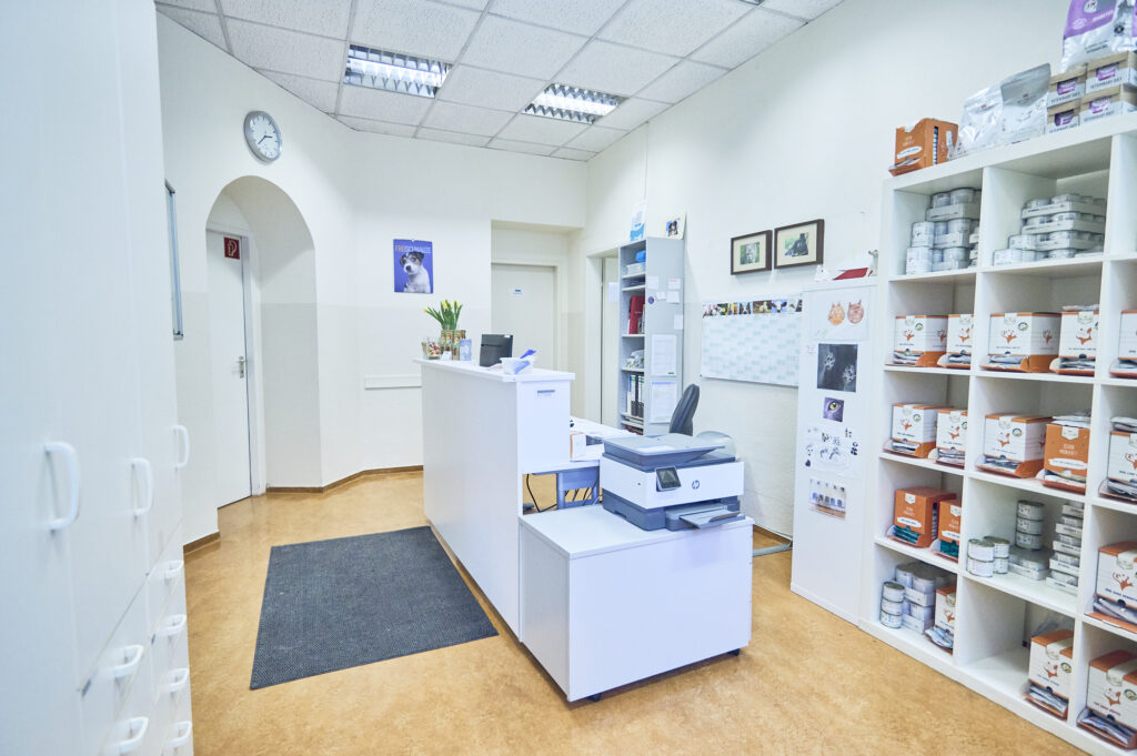 Empfangsbereich einer Tierarztpraxis mit weißer Rezeption, Regalen voller Tierfutter und Produkte, Drucker, Wanduhr und hellem Innenraum.